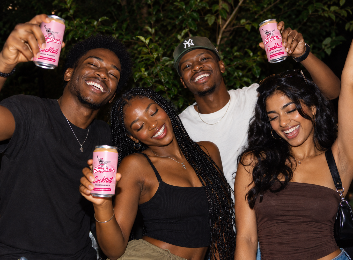 Three friends laughing at night, each holding up a pink Brightside Mango Margarita can.